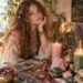 Beautiful woman with an ethereal bohemian appearance sitting beside a magical altar that includes rose quartz, green aventurine, tarot cards, pink and green candles, dried roses, lavender, and a small Libra zodiac symbol
